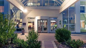 Entrada de hotel com átrio de vidro, placa com 'Mercure' e calçada pavimentada com vegetação.