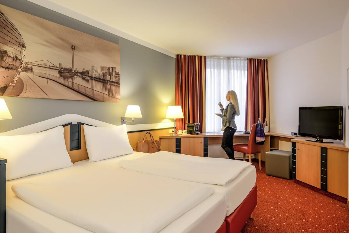 A hotel room with a large bed, a desk with a TV, a window with curtains, and a cityscape painting on the wall.