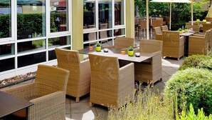 Área de terraço ao ar livre com móveis de vime e guarda-sóis, cercada por vegetação e um edifício com janelas amplas.