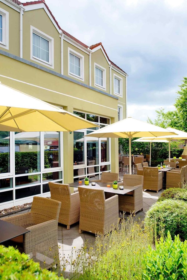 An outdoor patio area with wicker furniture and umbrellas, surrounded by greenery and a building with large windows. - Mercure Hotel Düsseldorf - Ratingen (Ratingen)