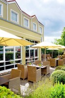 An outdoor patio area with wicker furniture and umbrellas, surrounded by greenery and a building with large windows.