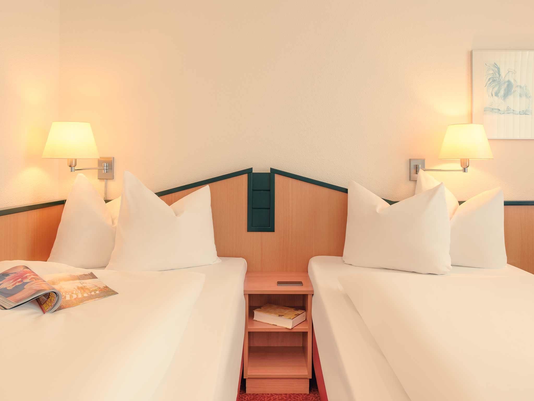 Habitación de hotel con dos camas, cada una con sábanas y almohadas blancas, y lámparas de pared a cada lado. Un mesita de noche de madera con un libro y un teléfono inteligente se encuentra entre las camas.