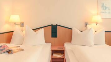Habitación de hotel con dos camas, cada una con sábanas y almohadas blancas, y lámparas de pared a cada lado. Un mesita de noche de madera con un libro y un teléfono inteligente se encuentra entre las camas.