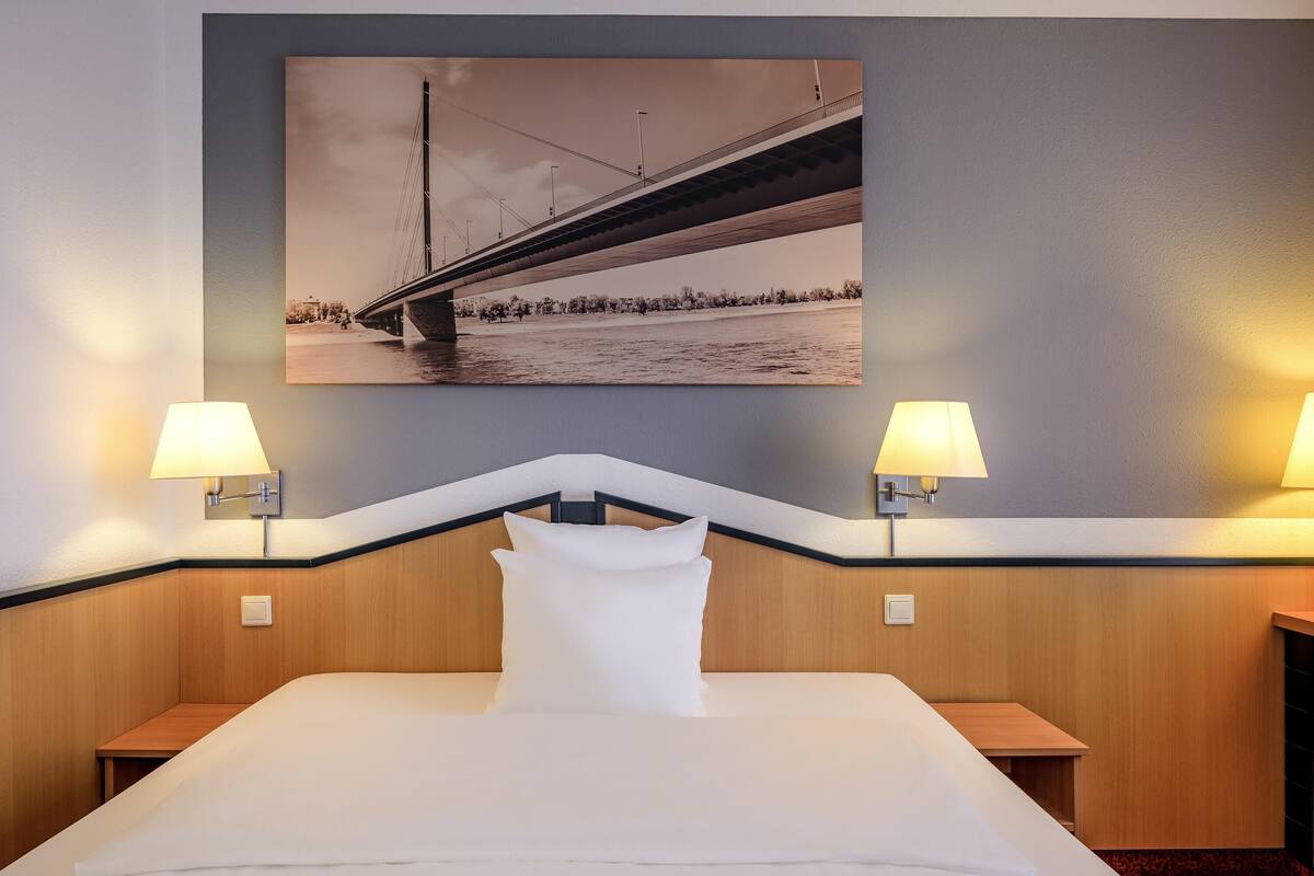 A hotel room with a bed, two bedside lamps, a headboard, a wall-mounted picture of a bridge, and a wooden headboard.