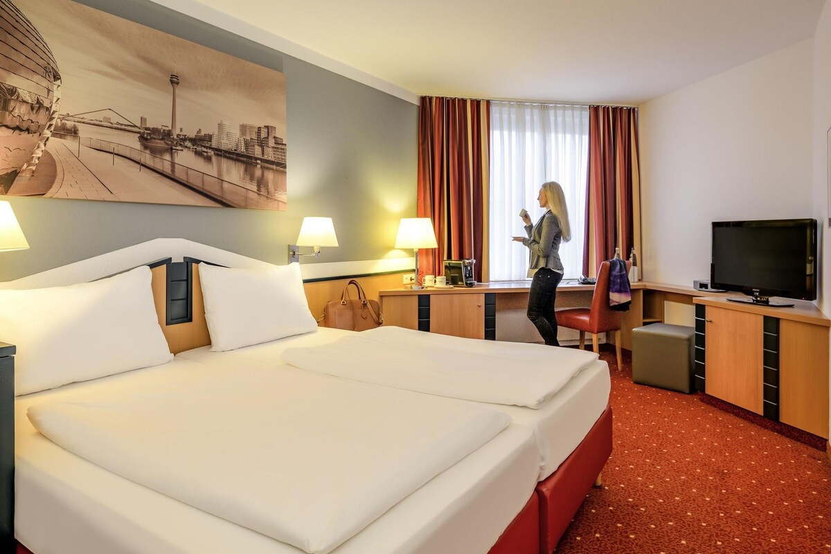 A hotel room with a large bed, a desk, a television, and a cityscape painting.