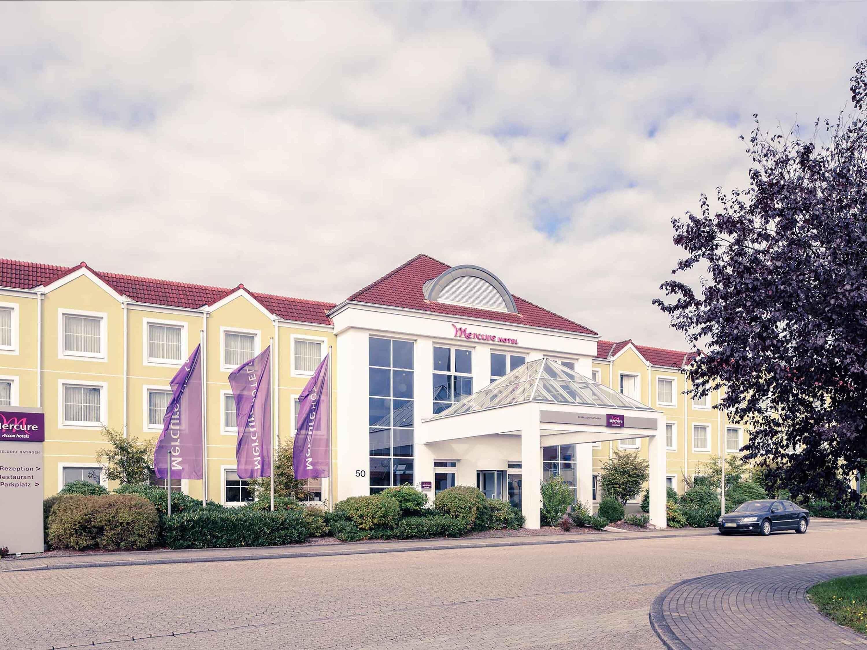 A hotel with a yellow facade, red roof, and a large glass entrance. There are flags and a sign for Mercure hotel.