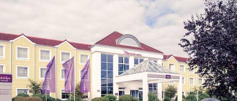 A hotel with a yellow facade, red roof, and a large glass entrance. There are flags and a sign for Mercure hotel.