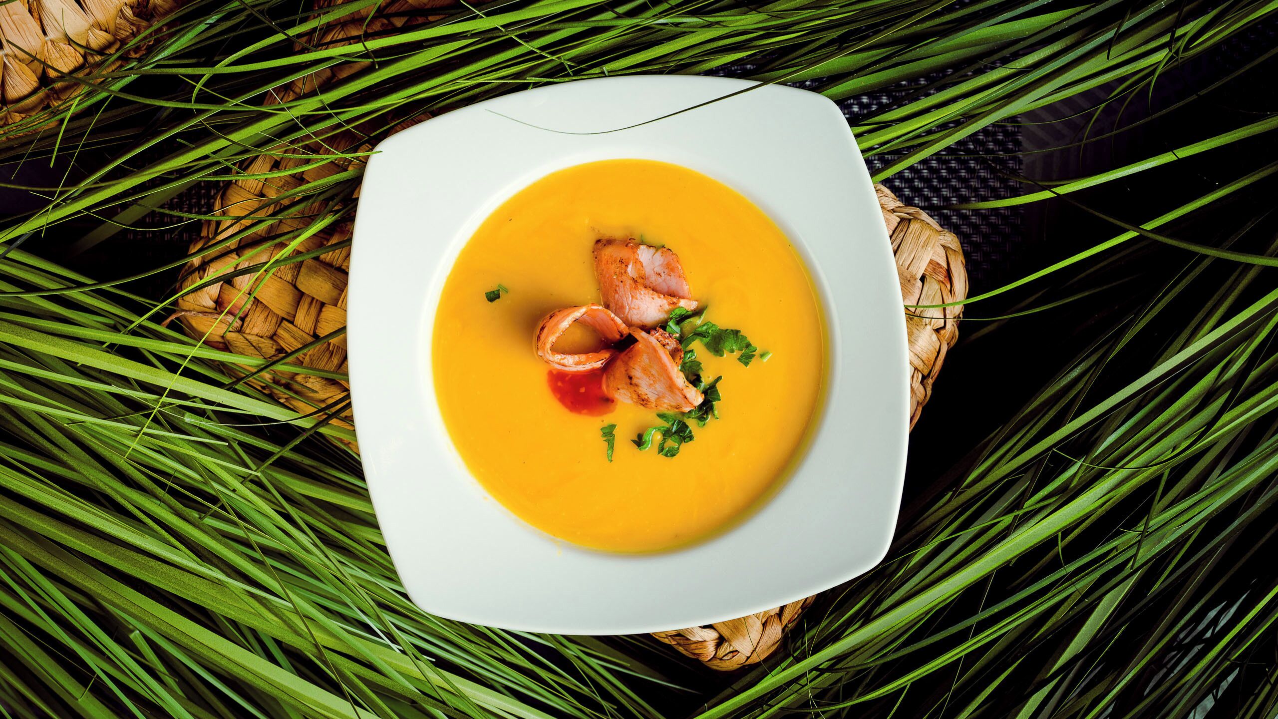 A white plate with a yellow soup and two pieces of meat, surrounded by green leaves.