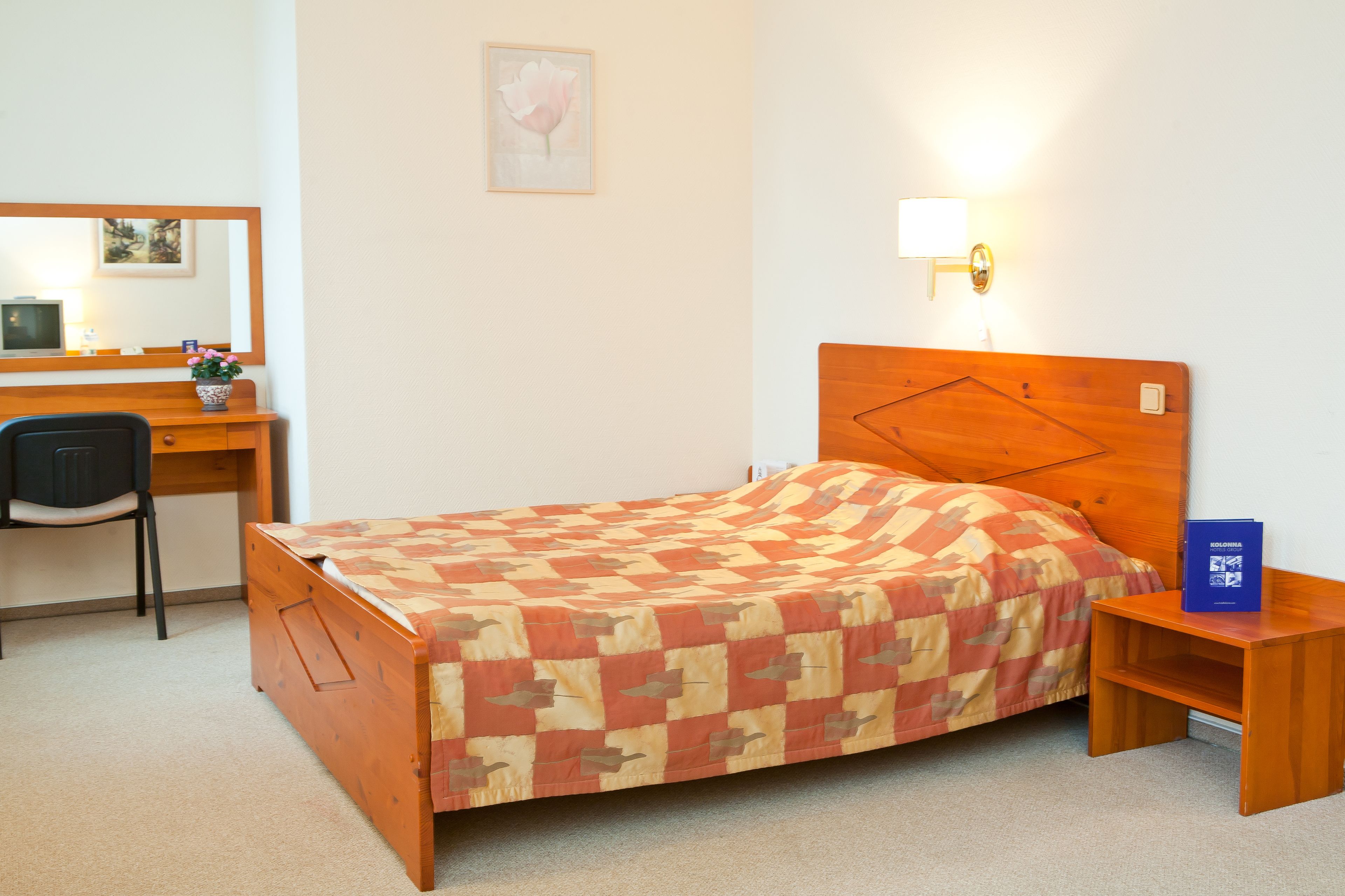 A bedroom with a wooden bed, a desk with a chair, a bedside table, and a wall-mounted lamp.