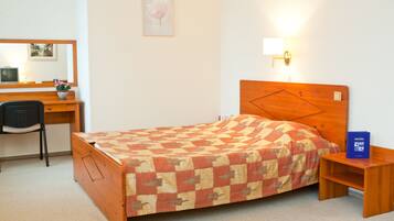 A bedroom with a wooden bed, a desk with a chair, a bedside table, and a wall-mounted lamp.