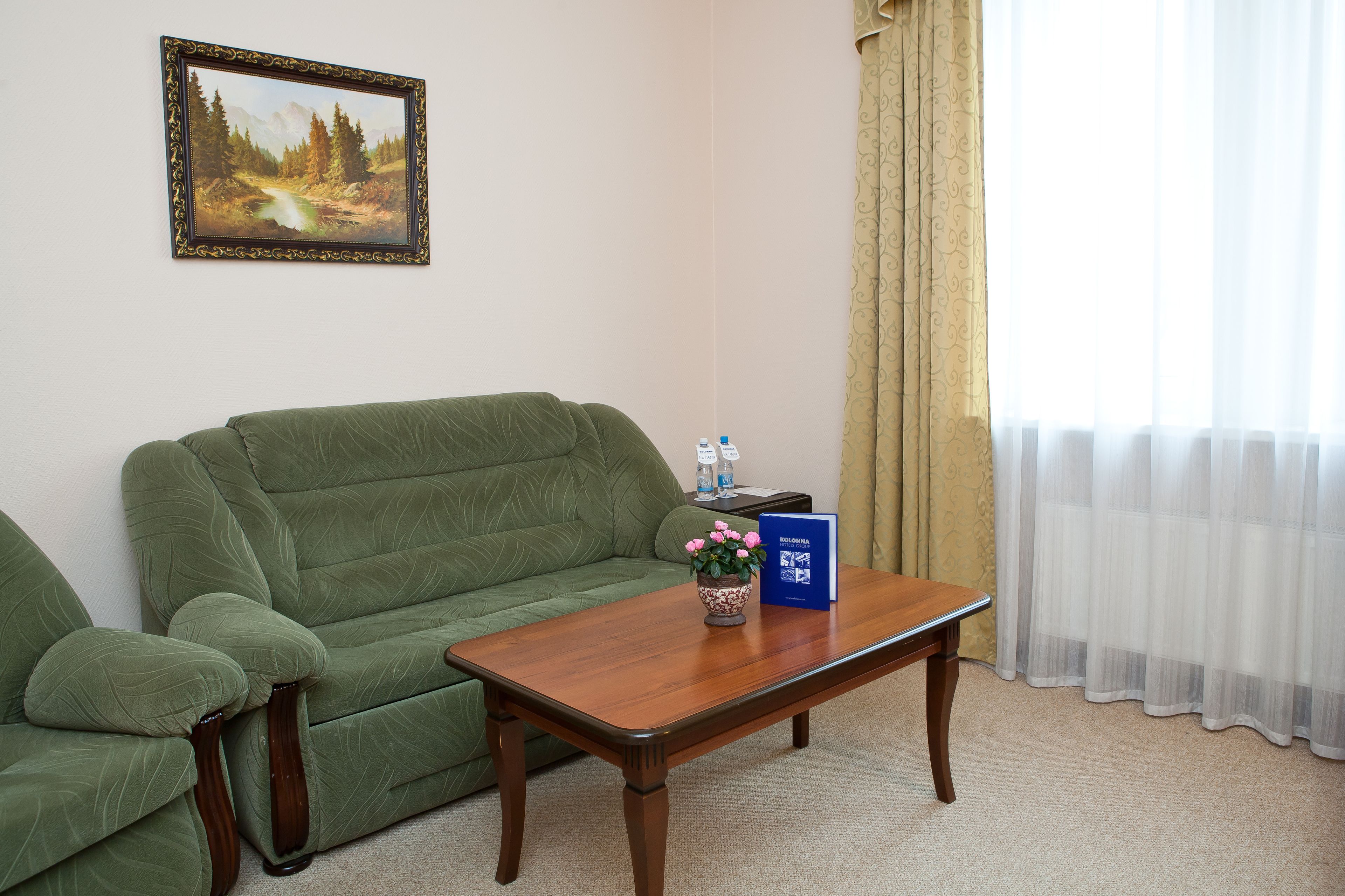 Una habitación con sofá verde, una mesa de centro de madera con un jarrón de flores rosas, un cuadro en la pared y una ventana con cortinas traslúcidas.