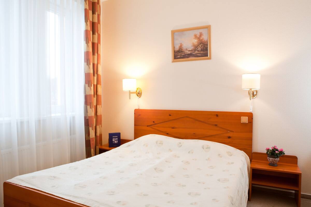 Een hotelkamer met een houten hoofdeinde, twee nachtkastjes, een bed met een bloemenprint en een schilderij aan de muur, plus een raam met dunne gordijnen.