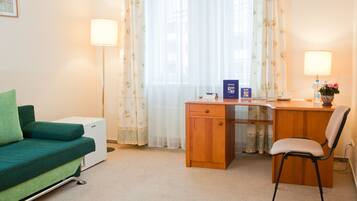 A hotel room with a green sofa, a wooden desk, a chair, and a small white appliance.