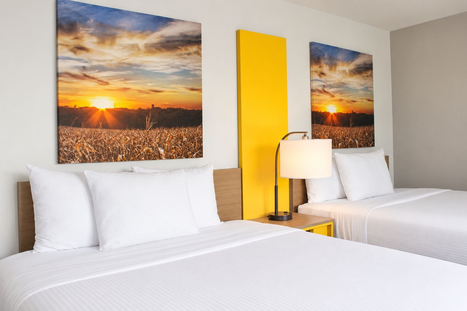 Une chambre d’hôtel avec deux lits, une table de chevet avec une lampe et deux grandes peintures représentant un coucher de soleil accrochées au mur.