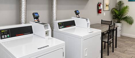 A laundry area with two Speed Queen commercial dryers, a dining table with chairs, and a potted plant.