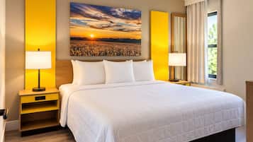 A hotel room with a large bed, two bedside tables with lamps, a nightstand, a large window with curtains, and a sunset painting on the wall.
