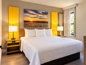 Habitación de hotel con una cama grande, dos mesitas de noche con lámparas, una mesita auxiliar, un amplio ventanal con cortinas y un cuadro de atardecer en la pared.