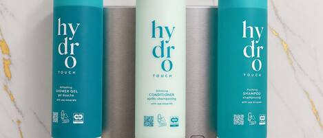 Trois flacons de produits de la gamme Hydra Touch : gel douche, après-shampoing et shampoing, chacun étant doté d’un code QR et de symboles écologiques.