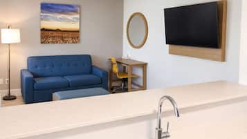 A modern kitchenette with a sink, a blue sofa, a small table with a chair, a wall-mounted TV, and a painting of a field.
