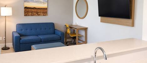 A modern kitchenette with a sink, a blue sofa, a small table with a chair, a wall-mounted TV, and a painting of a field.
