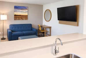 Una kitchenette moderna con un lavamanos, un sofá azul, una mesita con una silla, un televisor de pared y un cuadro de un campo.