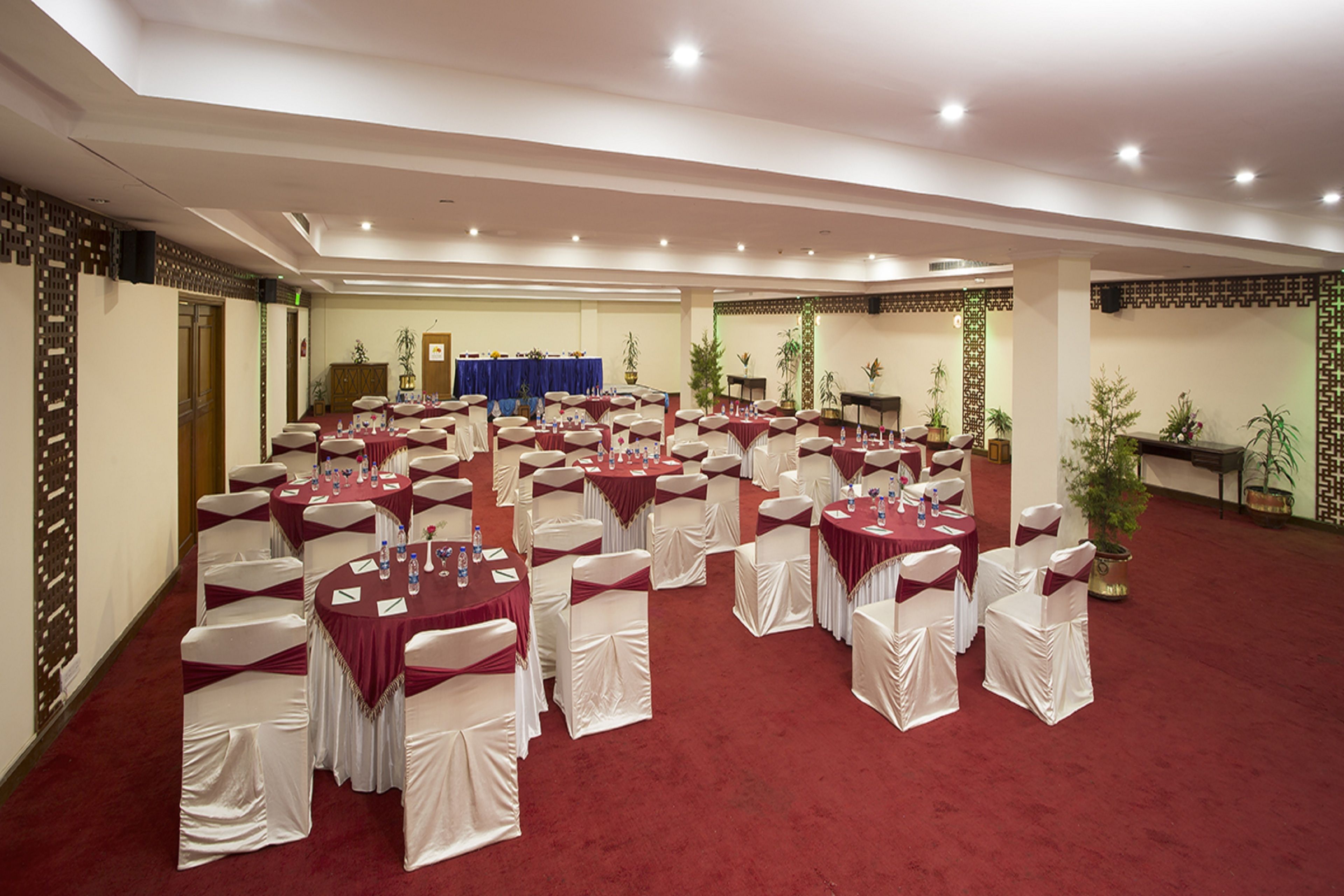 A banquet hall with round tables and white chairs, a stage with a blue backdrop, and potted plants.