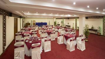 A banquet hall with round tables and white chairs, a stage with a blue backdrop, and potted plants.
