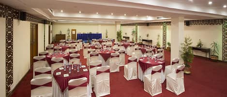A banquet hall with round tables and white chairs, a stage with a blue backdrop, and potted plants.