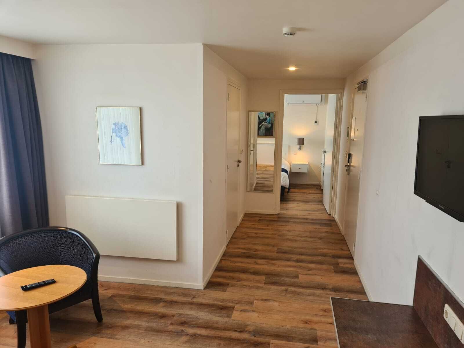 Een hotelgang met houten vloer, een bed op de achtergrond, een tv aan de muur en een schilderij.