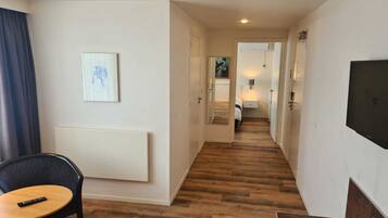 A hotel corridor with wooden flooring, a bed in the background, a TV mounted on the wall, and a painting on the wall.