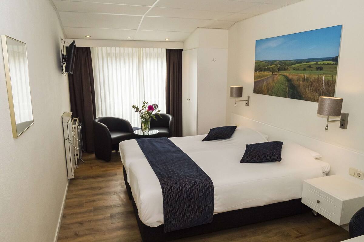 Une chambre d’hôtel avec un grand lit, un bureau, une chaise, une petite table ornée d’un vase de fleurs et un tableau représentant un paysage accroché au mur.