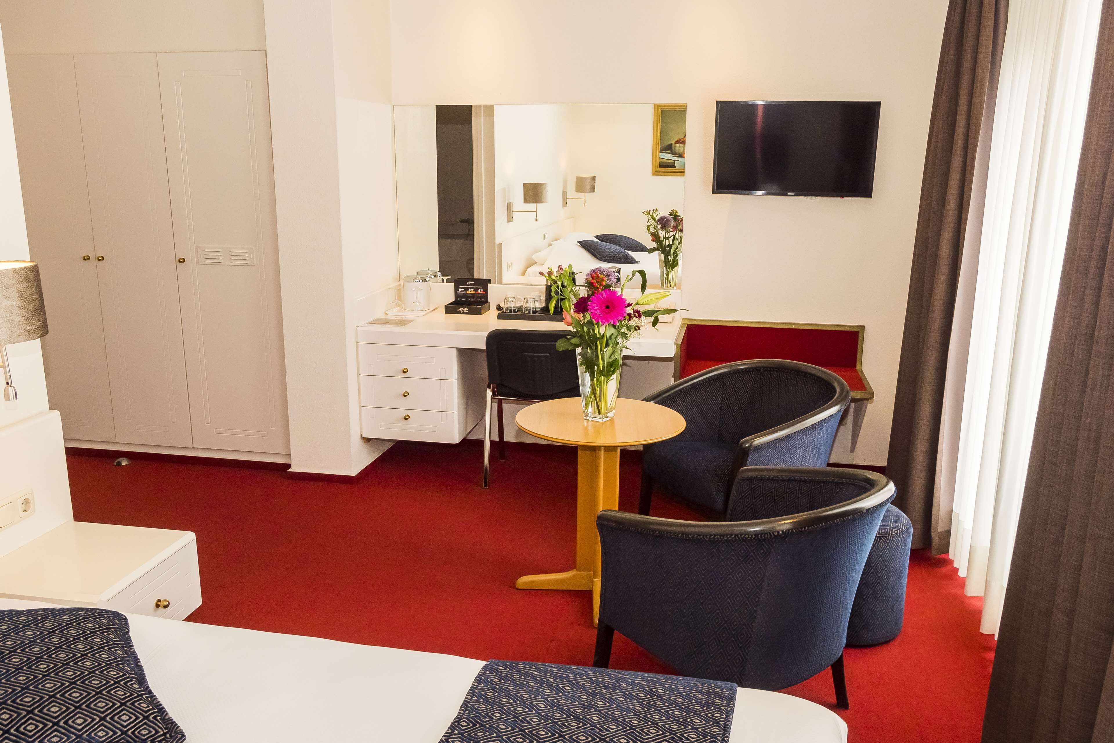 Een hotelkamer met een bed, een bureau met stoel, een bijzettafeltje met bloevasen, een aan de muur bevestigde televisie en een kledingkast.