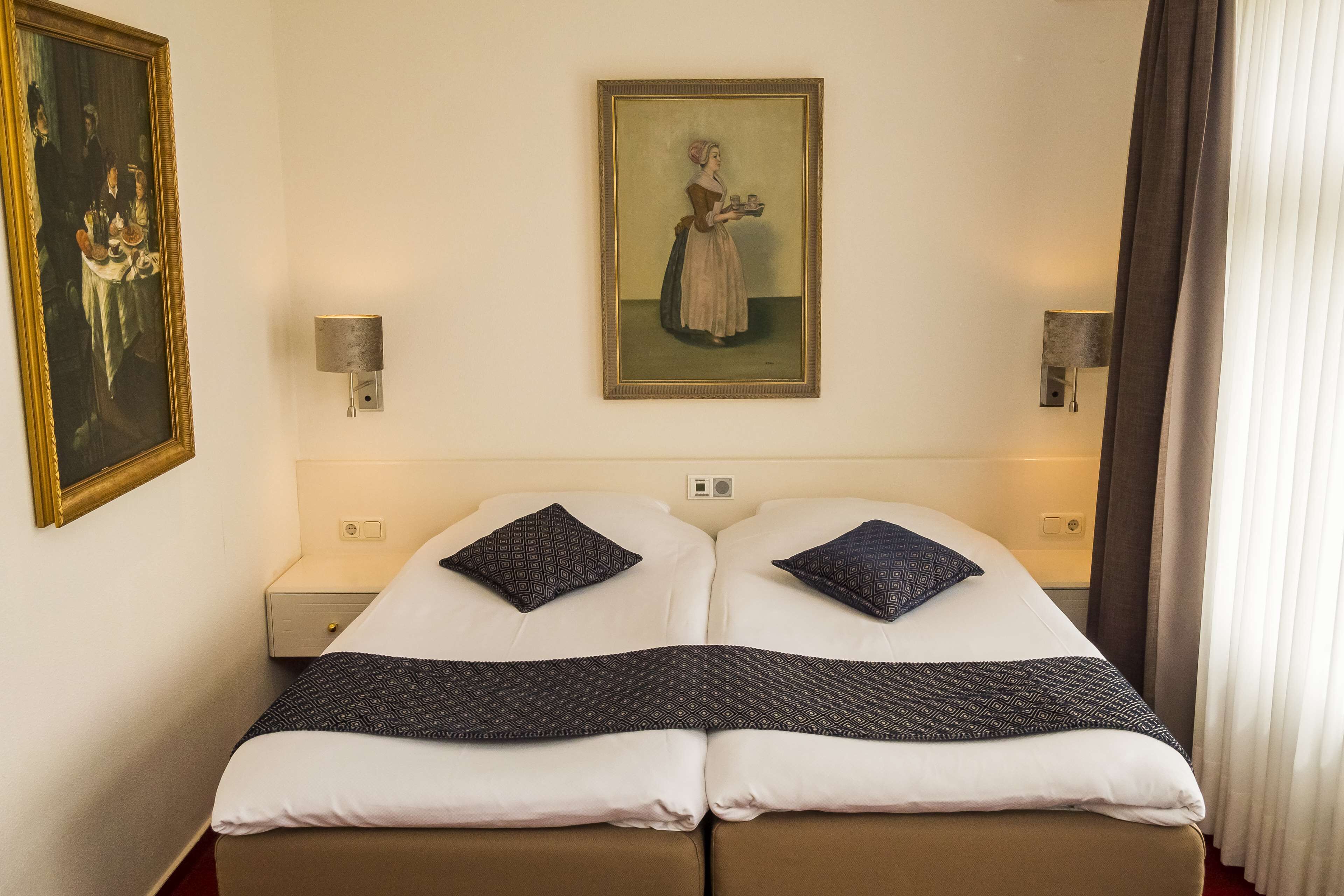 A double bed with two pillows, a painting on the wall, and a nightstand with a lamp.