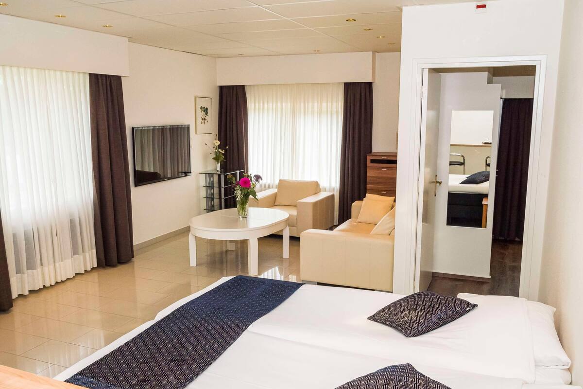Une chambre d’hôtel comprenant un lit, un coin salon avec une chaise et une table, une télévision et donnant sur le couloir.