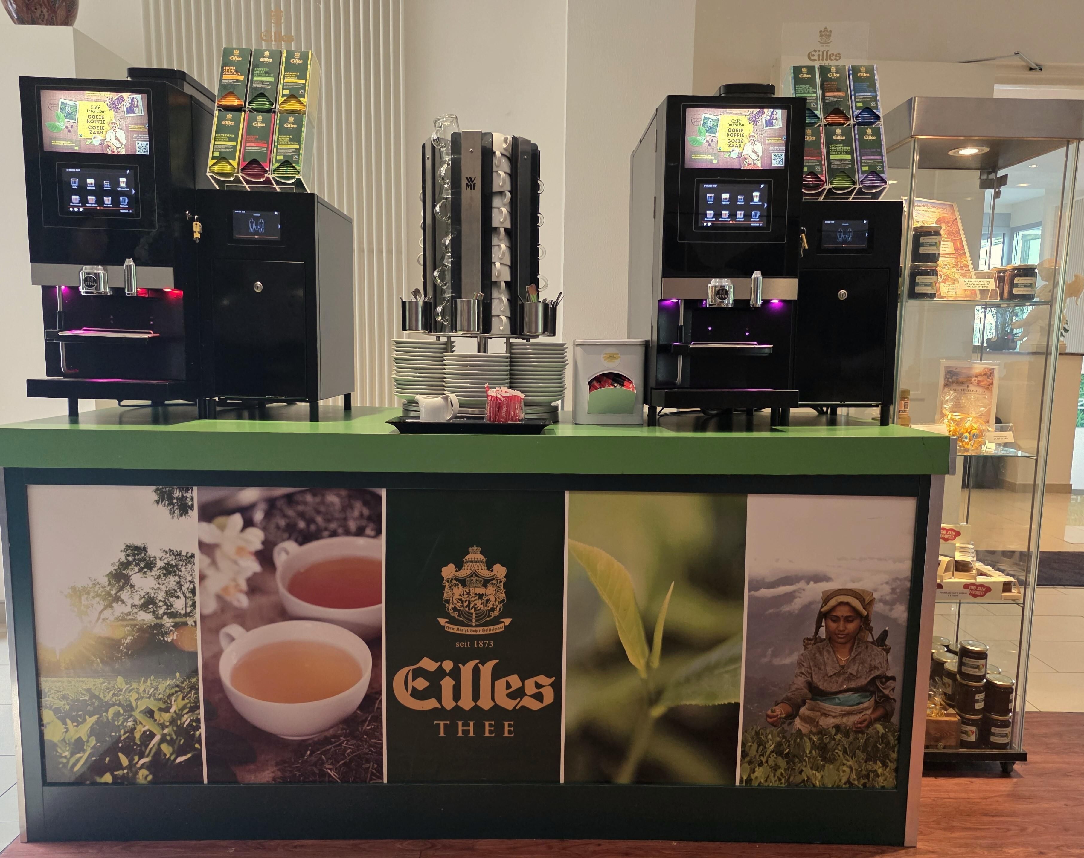 A coffee station with two espresso machines, a display of coffee products, and a counter with the brand 'Eilles Thee' prominently displayed.