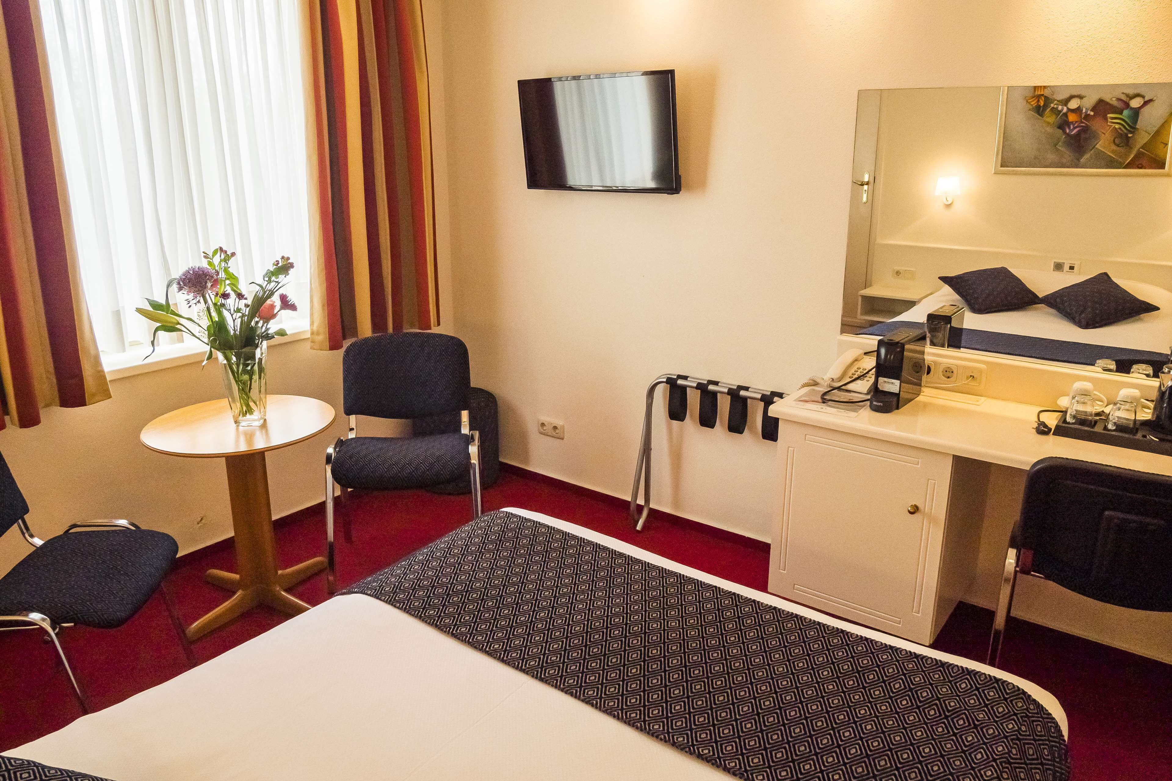 A hotel room with a bed, a desk, a chair, a small table with a vase of flowers, a television, and a painting on the wall.
