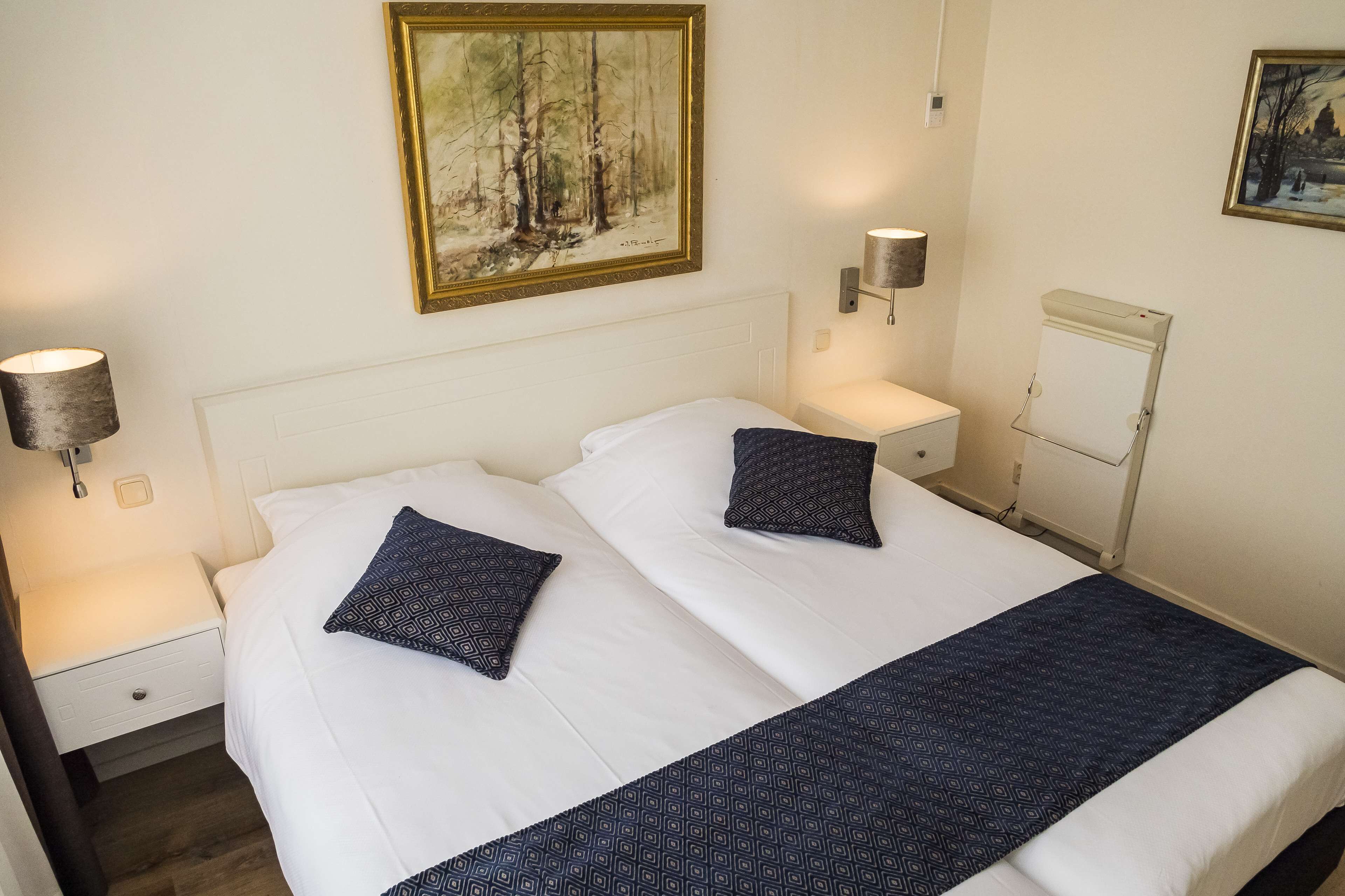 Een tweepersoonsbed met witte lakens en twee gestreepte kussens, een nachtkastje met een lamp, en een wandplank.
