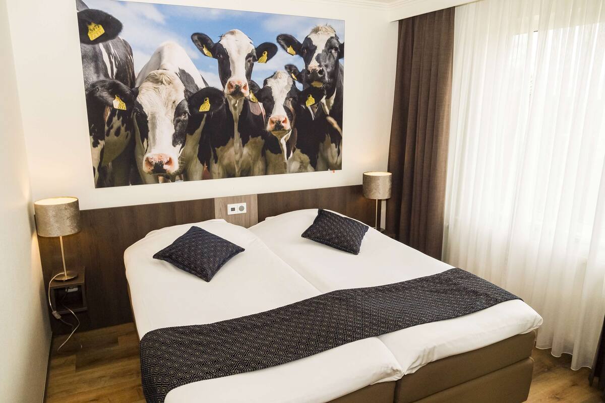 Une chambre d’hôtel avec un grand lit, deux oreillers, une lampe de chevet et un tableau représentant des vaches accroché au mur.
