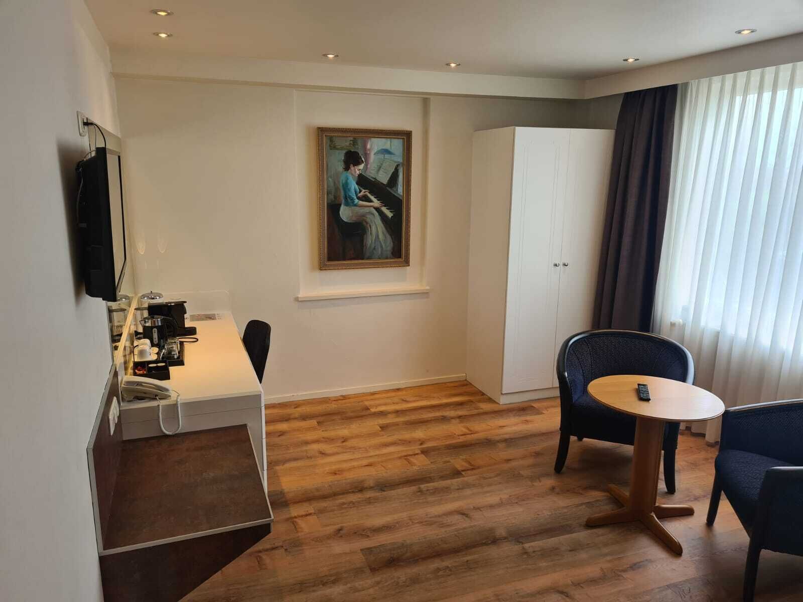 A hotel room with a wooden floor, a desk with a telephone and coffee machine, a chair, a small round table, a wardrobe, and a painting on the wall.