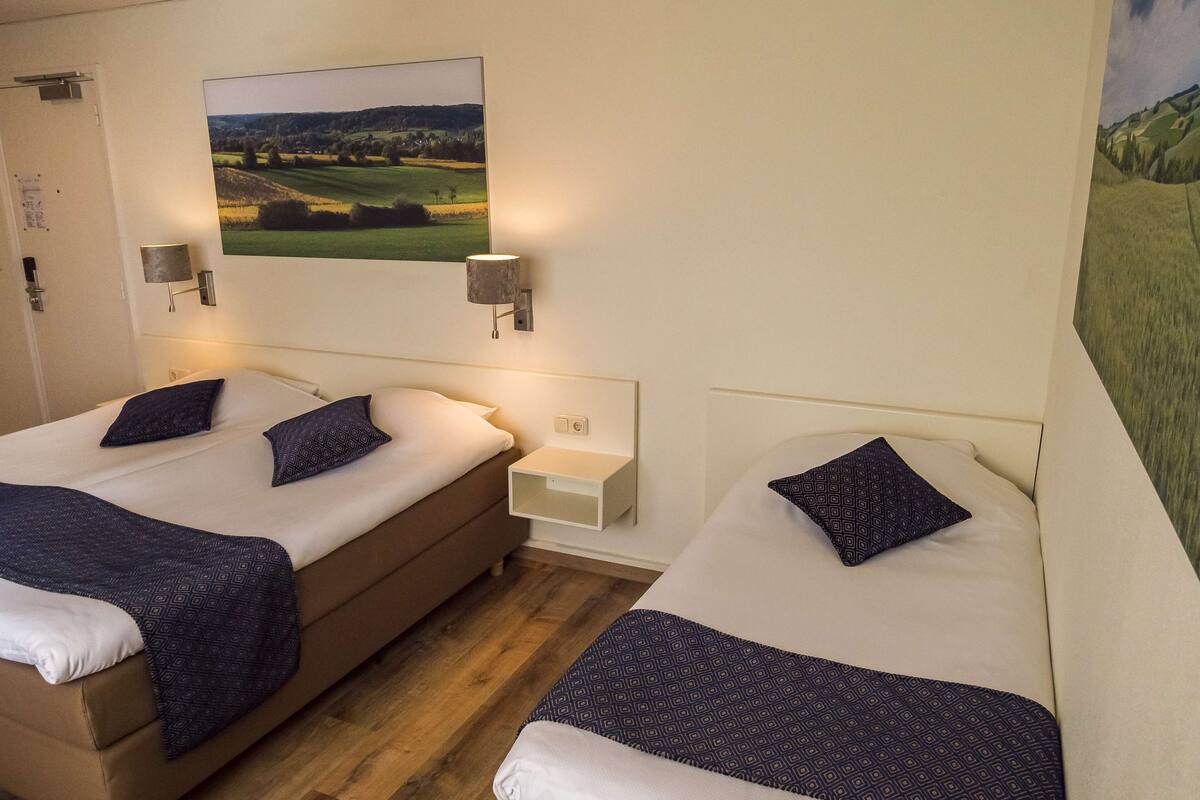 Une chambre d’hôtel avec deux lits, une table de chevet, une lampe et un tableau représentant un paysage.