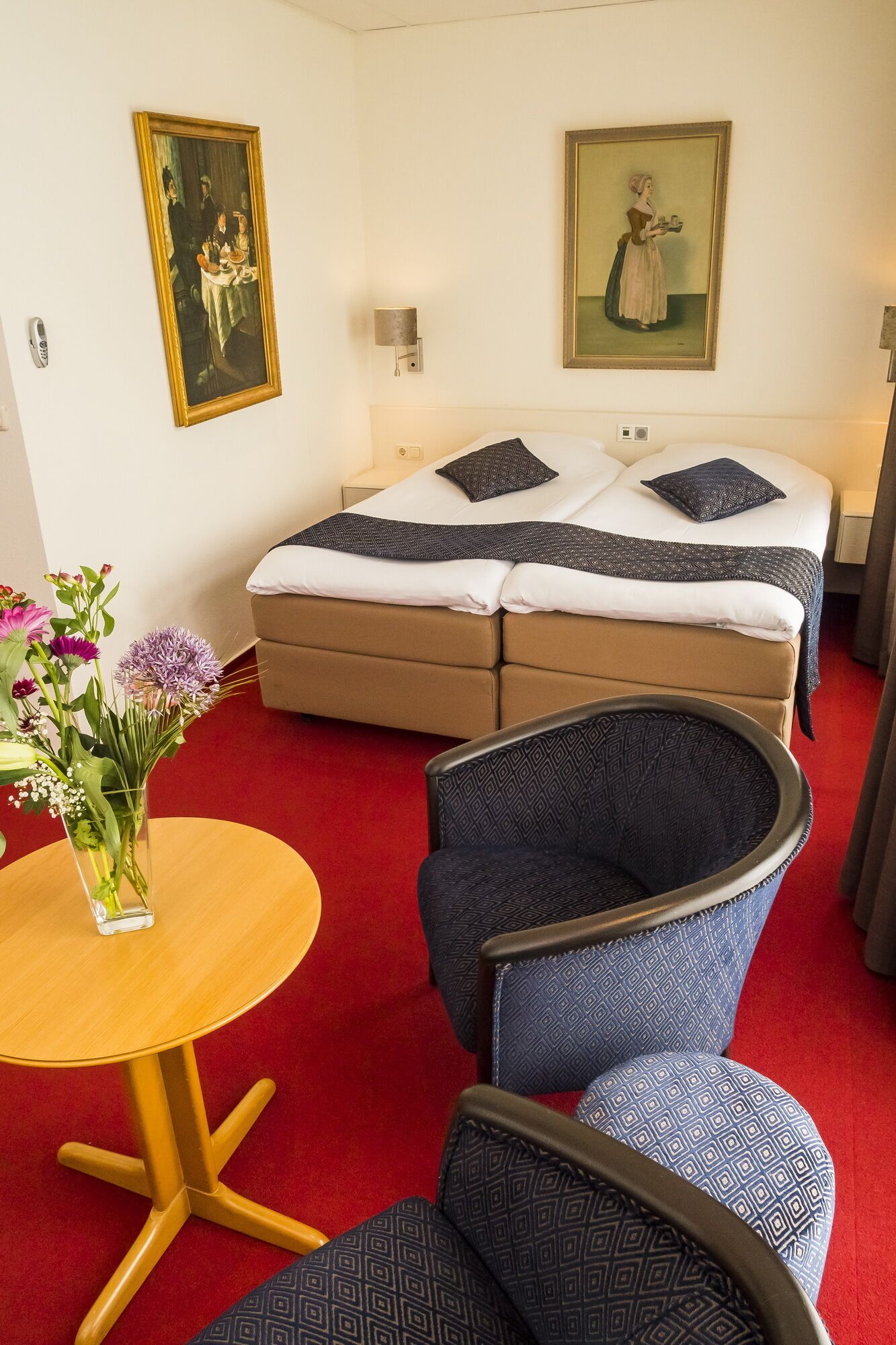 A hotel room with a bed, two bedside tables, a small round table with a vase of flowers, and two patterned chairs.