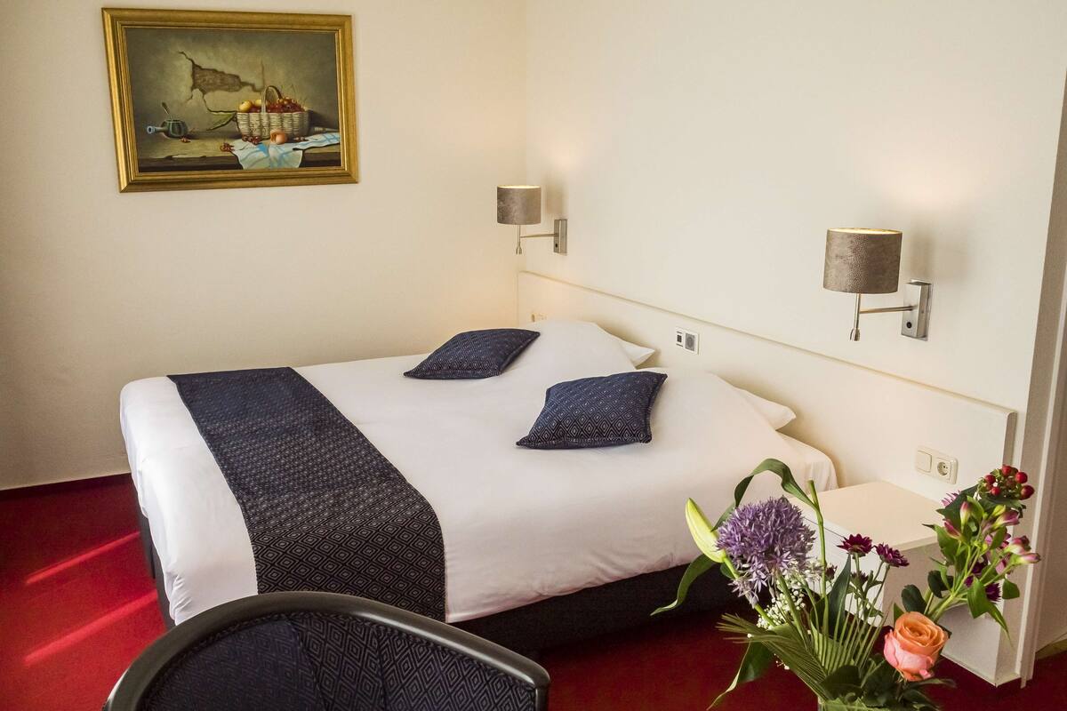 Une chambre d’hôtel avec un lit, deux lampes fixées au mur, un tableau et une petite table ornée de fleurs.