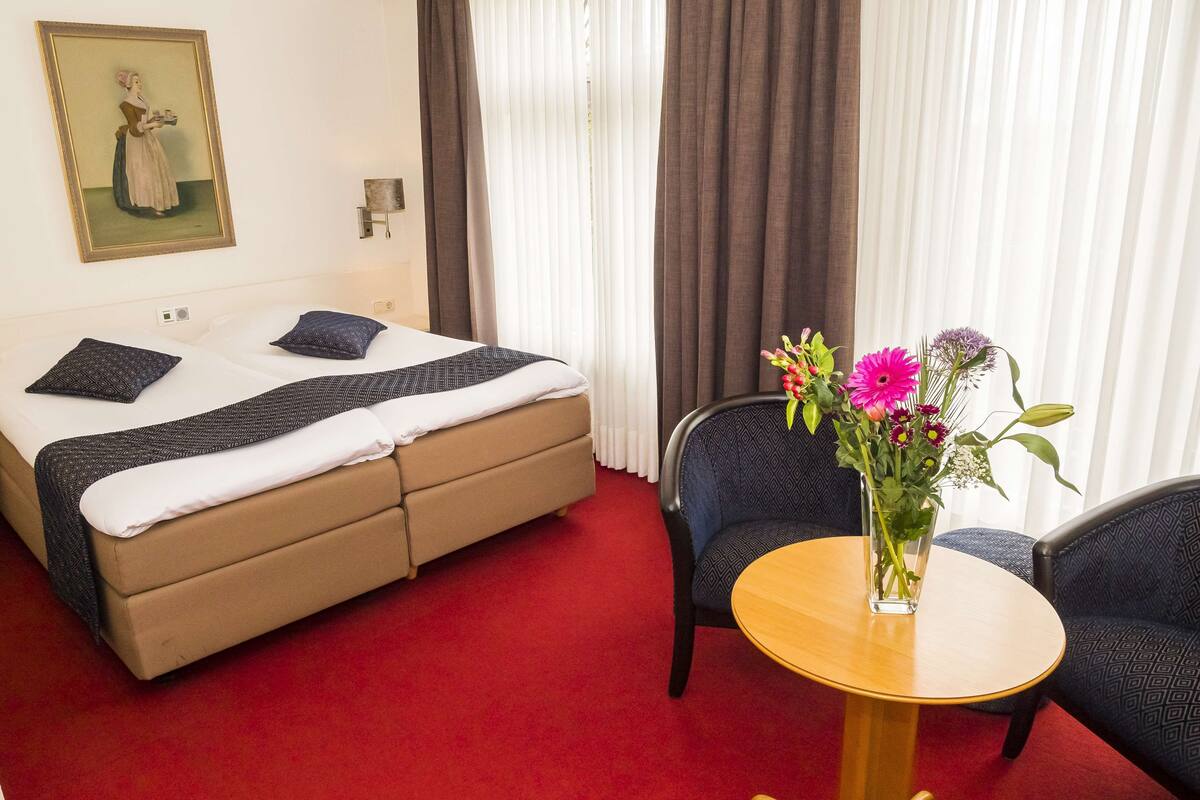 Une chambre d’hôtel avec un lit, une petite table ronde ornée d’un vase de fleurs, une chaise et un tableau accroché au mur.