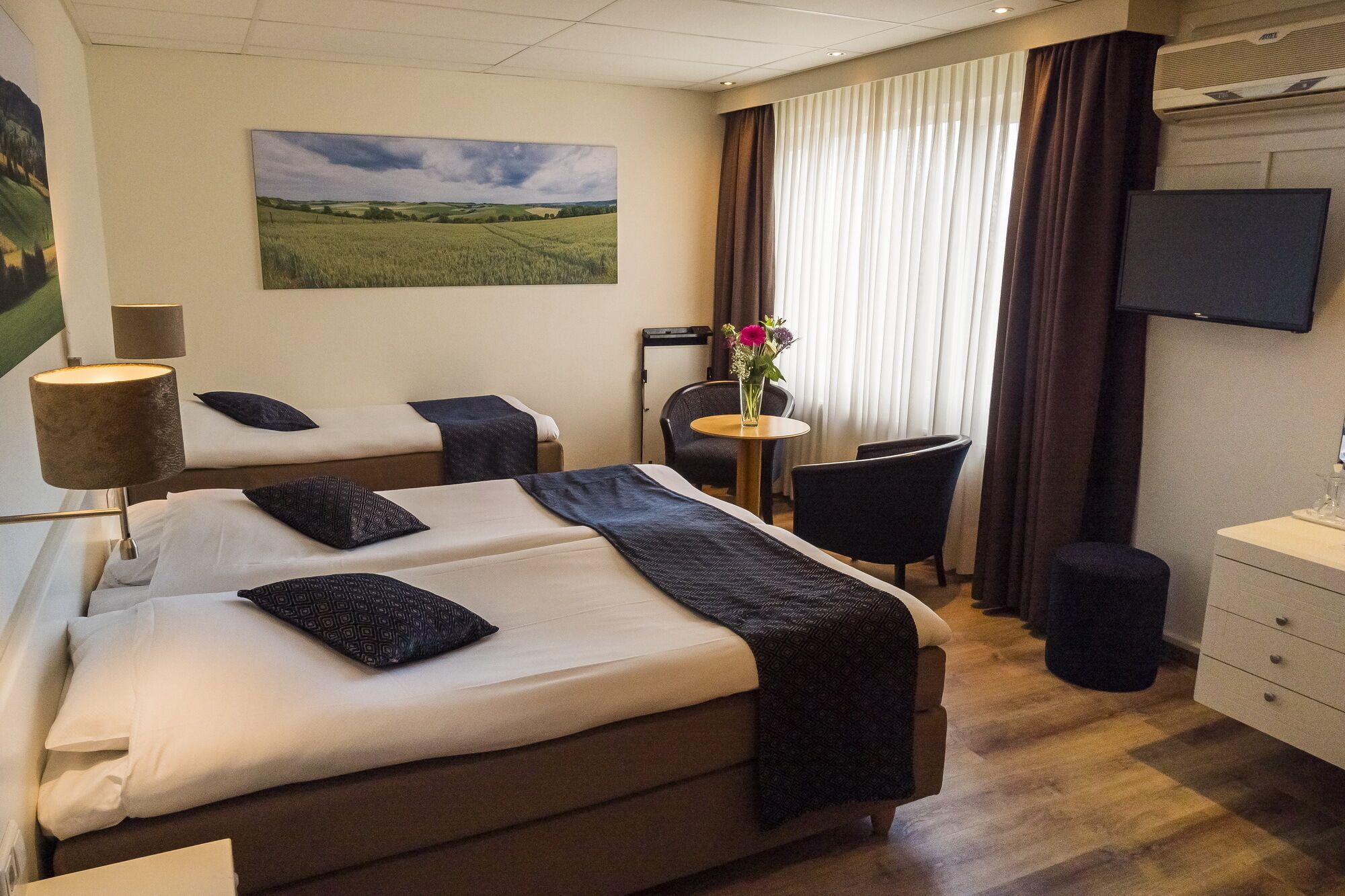Ett hotellrum med två sängar, ett skrivbord, en stol, en tv och en tavla som föreställer ett landskap.