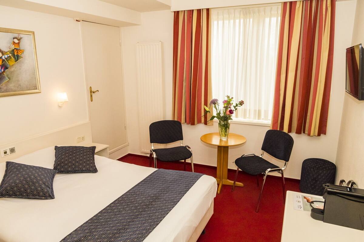 Une chambre d’hôtel avec un grand lit, deux chaises, une petite table et une fenêtre avec des rideaux à rayures.