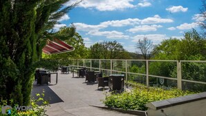 Een terras buiten met tafels en stoelen, omgeven door groen en een heldere lucht.