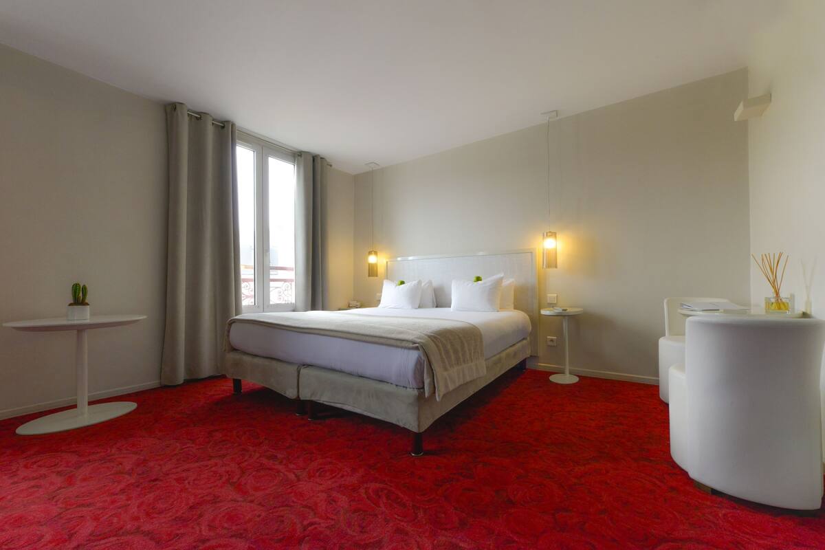 A hotel room with a large bed, a small table, a chair, and a decorative vase.