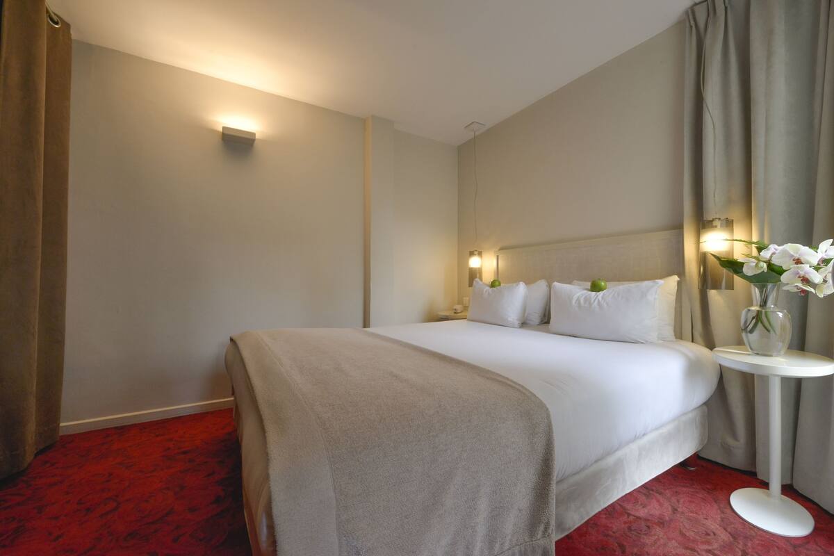 A hotel room with a large bed, a bedside table with a vase of flowers, and a wall-mounted light fixture.