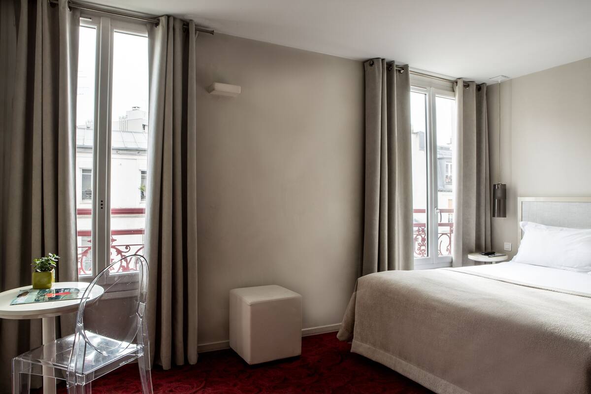 A hotel room with a bed, a small table with a chair, a nightstand, and a potted plant.