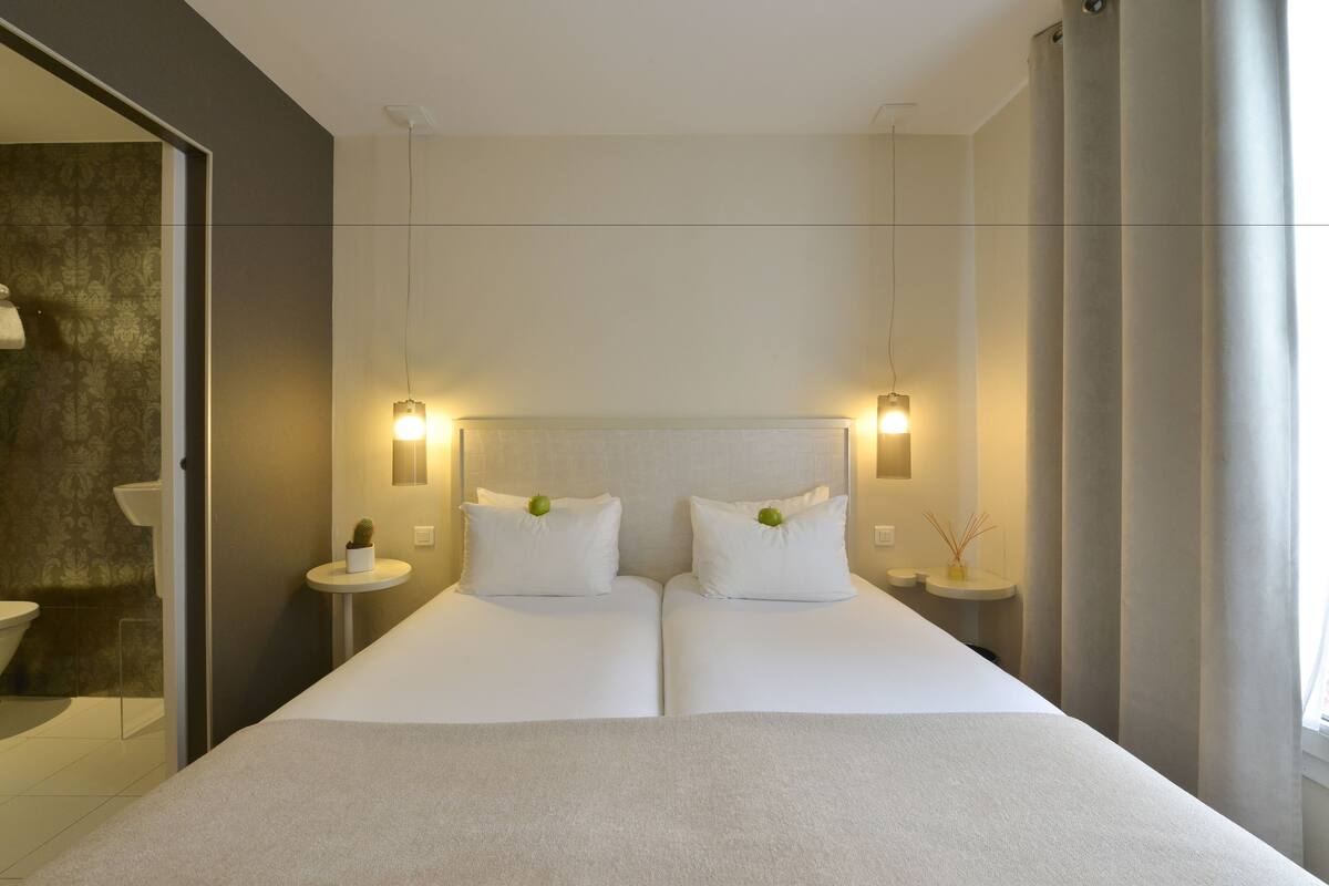 A modern hotel room with a large bed, two bedside tables, a small round table, and a mirror.